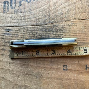 Puede incluir: Un cuchillo de bolsillo cerrado con mango de lat&oacute;n y hoja plateada, que mide aproximadamente 10 cm de largo. El cuchillo est&aacute; colocado junto a una regla de madera marcada en pulgadas, con el filo de la hoja hacia arriba.