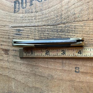 Puede incluir: Un cuchillo de bolsillo cerrado con mango de lat&oacute;n y hoja negra, junto a una regla de madera. La regla mide el cuchillo en aproximadamente 10 cm de largo.