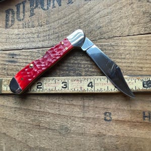 May include: A folding pocket knife with a textured red handle and a silver metal blade, positioned against a yellow measuring tape that shows the knife measures approximately 3.5 inches in length when partially opened.