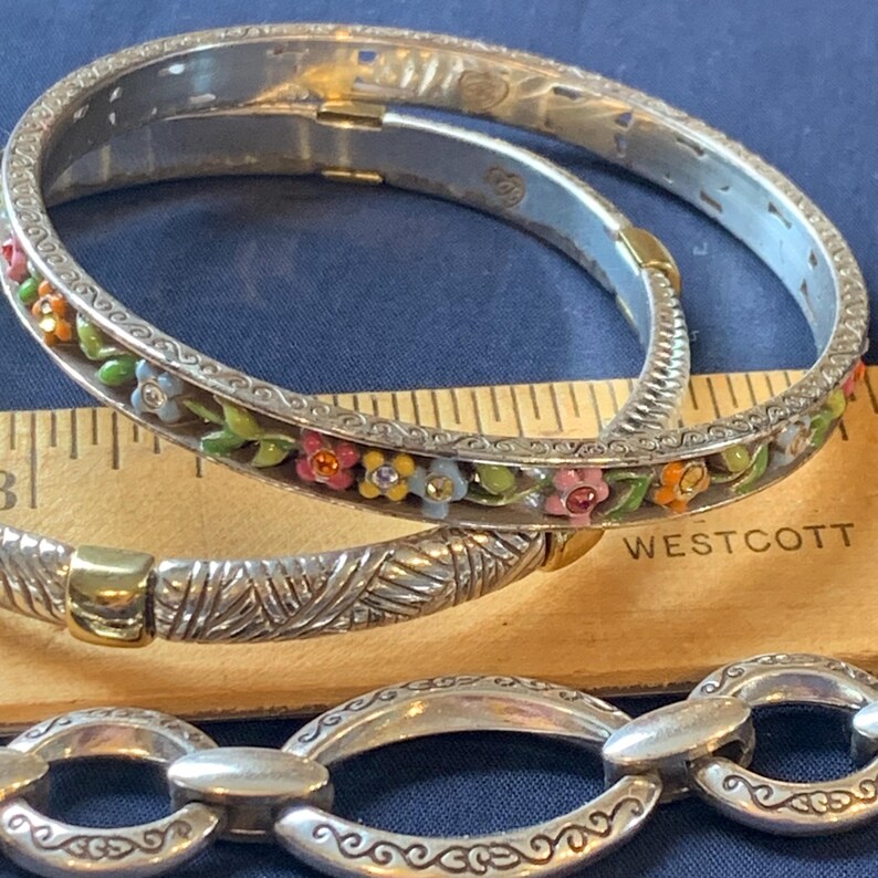 May include: Silver-tone bracelets with floral and decorative designs. One bracelet features colorful enamel flowers and leaves. A ruler is placed next to the bracelets. The bracelets are on a blue surface.
