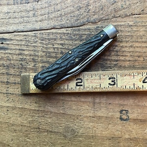 May include: A vintage pocket knife with a dark, textured handle and a silver-colored bolster. The blade is partially open, revealing a shiny metal edge. A ruler is placed next to the knife, showing a measurement of approximately 3 inches.