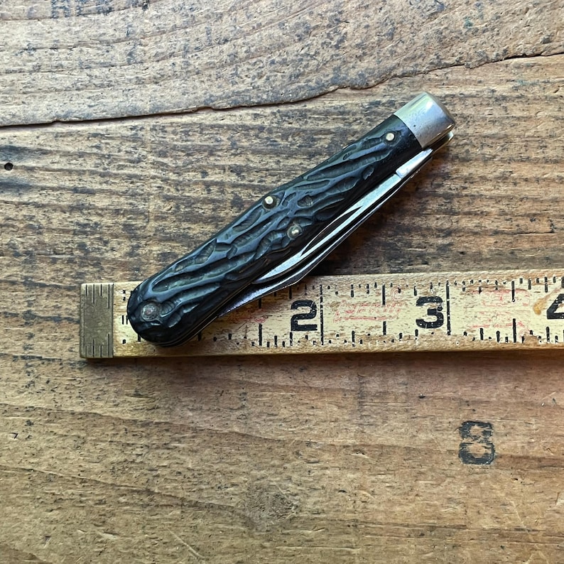 May include: A vintage pocket knife with a dark, textured handle and a silver-colored bolster. The blade is partially open, revealing a shiny metal edge. A ruler is placed next to the knife, showing a measurement of approximately 3 inches.