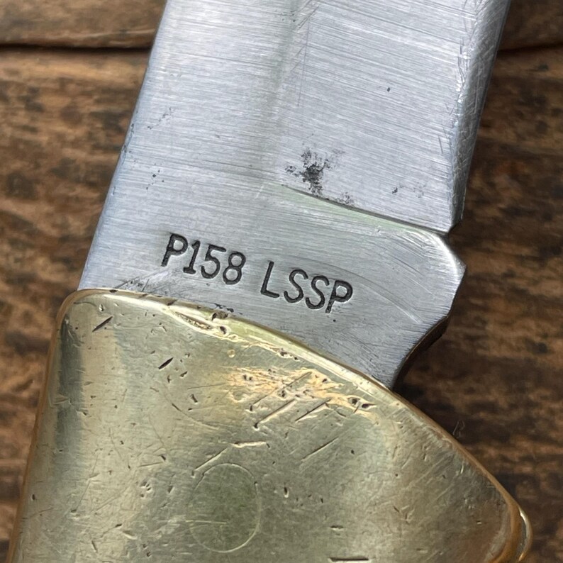 Puede incluir: Primer plano de una navaja plegable parcialmente abierta. La hoja plateada tiene el texto "P158 LSSP" grabado. El mango es dorado. El cuchillo est&aacute; sobre una superficie de madera.