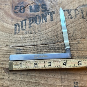 May include: A vintage pocket knife with a silver blade and handle, partially open. The handle has the text "SunKist" printed on it. A wooden ruler with inch measurements is next to the knife.