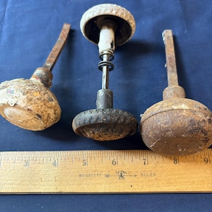 May include: Three vintage metal door knobs with attached square spindles, positioned above a wooden Westcott ruler. The knobs exhibit varying degrees of oxidation, textured surfaces, and weathered finishes in shades of brown, gray, and off-white.
