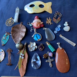 May include: A collection of various pendants and charms, including a leaf, a fish-shaped pendant, a crystal, a cross, and a gnome. The pendants are in various colors and materials, such as copper, silver, and gemstones.