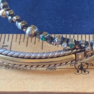 May include: Close-up of a bracelet with a silver and gold band. The top band is adorned with small, clear, and green gemstones. The bracelet is resting on a wooden ruler with inch markings, against a blue background.