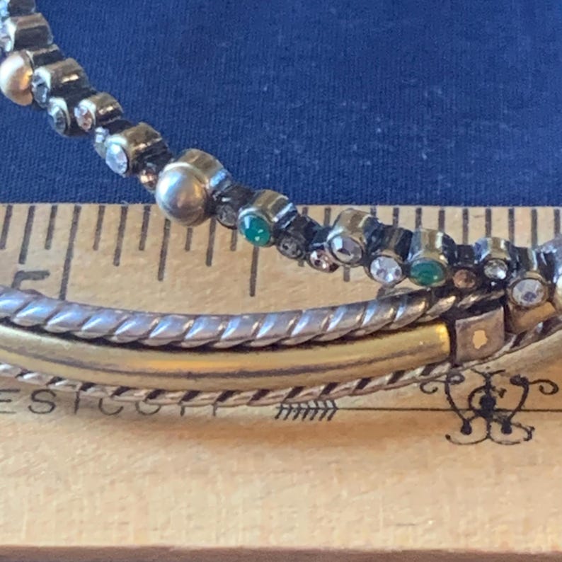May include: Close-up of a bracelet with a silver and gold band. The top band is adorned with small, clear, and green gemstones. The bracelet is resting on a wooden ruler with inch markings, against a blue background.