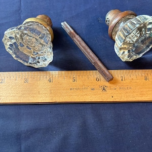 May include: Two clear glass door knobs with faceted surfaces and brass-toned metal bases, accompanied by a long, thin, threaded metal spindle. A wooden Westcott ruler is positioned below the items, showing the spindle length is about 3 inches.