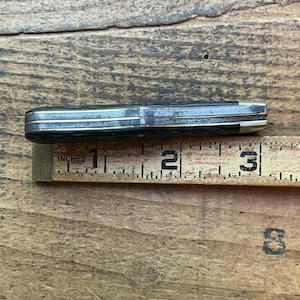 May include: A closed pocket knife with a black handle and silver accents, measuring approximately 3 inches (7.6 cm) in length. The knife is placed next to a wooden ruler with the word "INCHES" printed on it.