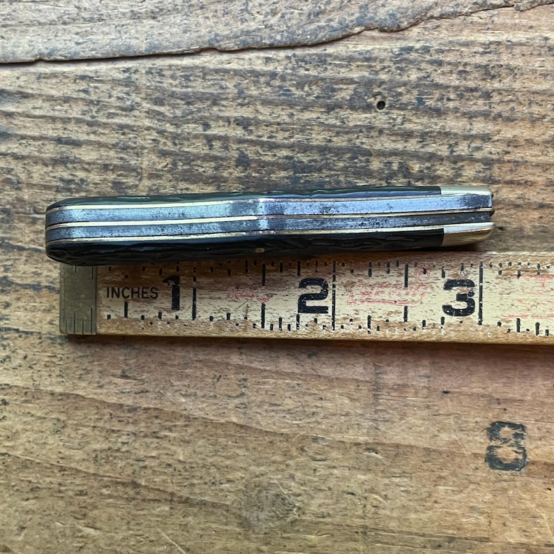 May include: A closed pocket knife with a black handle and silver accents, measuring approximately 3 inches (7.6 cm) in length. The knife is placed next to a wooden ruler with the word "INCHES" printed on it.