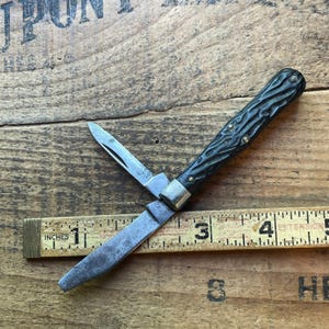 May include: A vintage pocket knife with a dark green textured handle and two blades, one partially open. The knife is positioned on a wooden surface next to a ruler marked in inches.