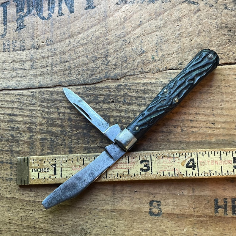 May include: A vintage pocket knife with a dark green textured handle and two blades, one partially open. The knife is positioned on a wooden surface next to a ruler marked in inches.