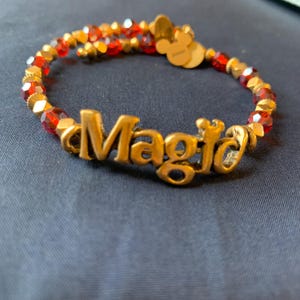 May include: A gold-toned bracelet with the word "Magic" in the center. The bracelet features red and gold beads, with a Mickey Mouse charm. The bracelet is on a dark blue surface.