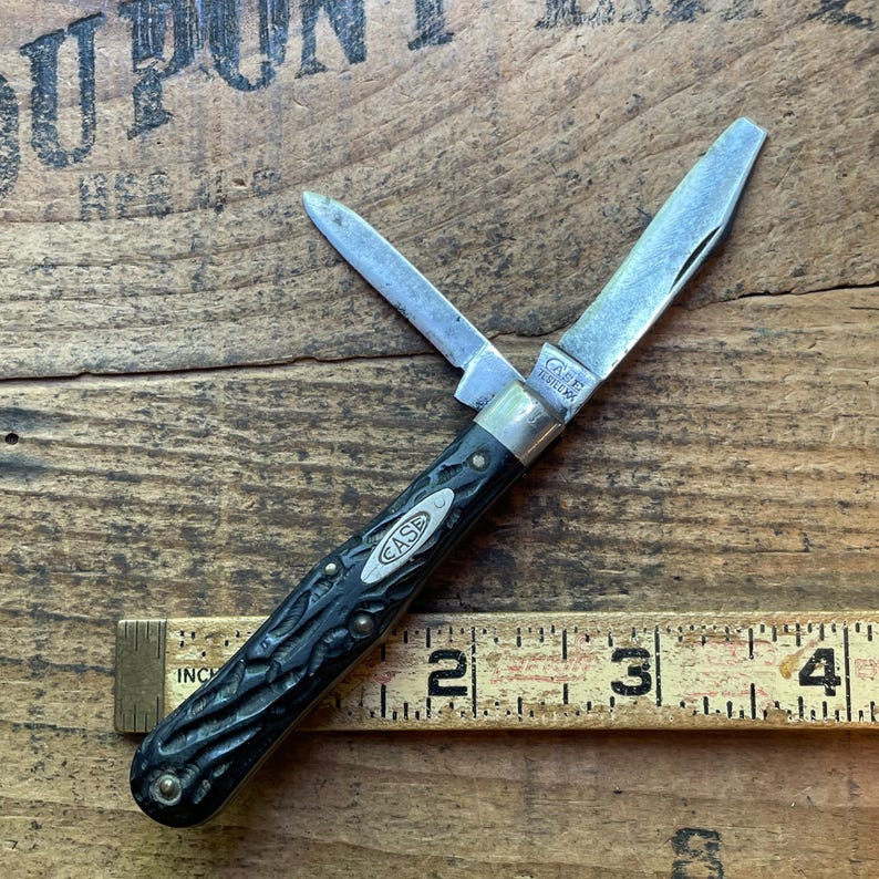 May include: A vintage Case pocket knife with two open blades. The knife has a black handle with a textured design and the Case logo. The blades are silver, and the knife is shown on a wooden surface with a ruler.