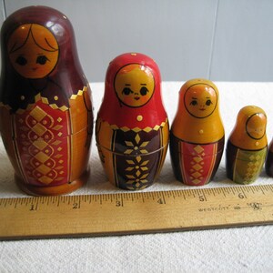 May include: A set of five nesting dolls, also known as Matryoshka dolls, in varying sizes. The dolls are painted with traditional designs in shades of red, brown, yellow, and gold. A wooden ruler is placed next to the dolls for size comparison.