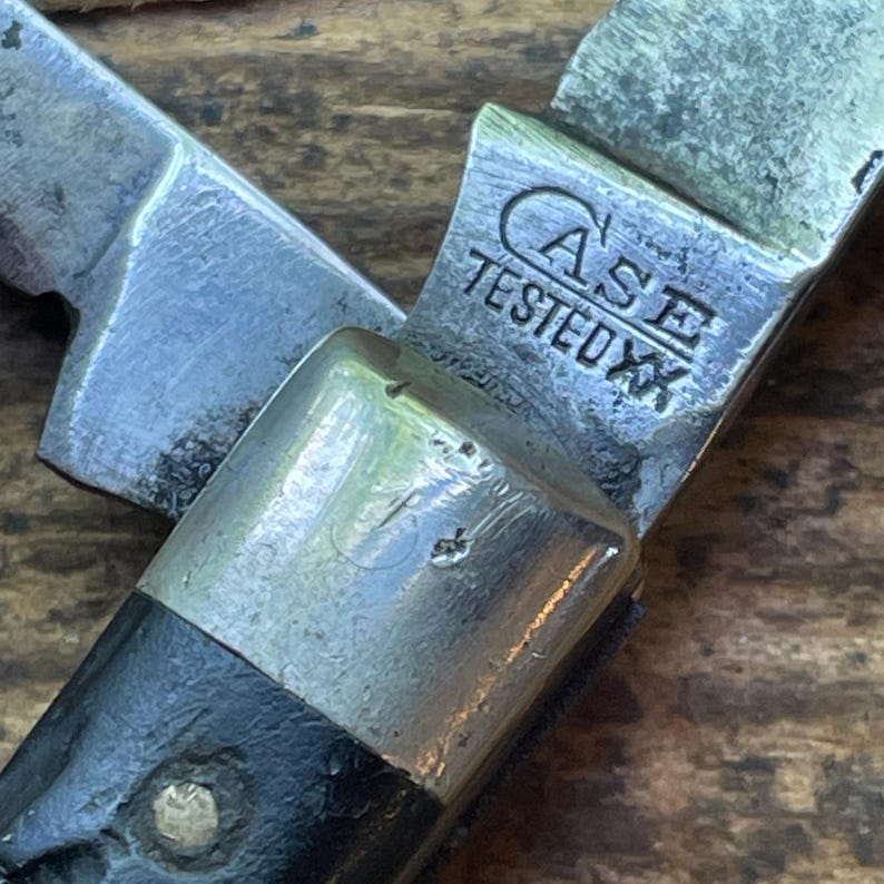 May include: Close-up of a vintage pocket knife with a partially open blade. The knife features a silver metal bolster and blade, with the text "CASE TESTED XX" etched on the metal. The handle is black. The knife is likely a collectible or a tool.