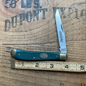 May include: A folding pocket knife with a dark green handle, brass bolsters, and a silver blade featuring the text SKOAL and SCHRADE USA. The knife is positioned above a wooden ruler showing a length of approximately 4.5 inches.
