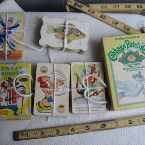 May include: A collection of vintage card games, including Crazy Eights, Cabbage Patch Kids, and a deck of fish cards. The cards are tied together with string and are displayed on a white surface.