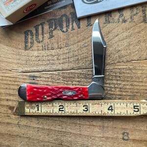 May include: A folding pocket knife with a bright red jigged bone handle and a single polished stainless steel blade. The knife is positioned against a yellow measuring tape, showing a closed length of approximately 3.5 inches.