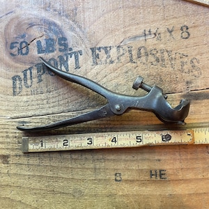 May include: An antique metal tool with curved handles and a screw adjustment. A wooden ruler with inch markings is placed next to the tool. The tool is set against a wooden surface with faded text.