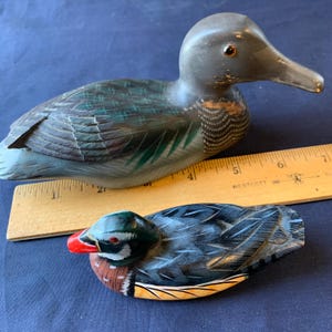 May include: Two wooden duck figurines. The larger duck is gray with green and black accents. The smaller duck has a red beak, brown chest, and a green head. A wooden ruler is placed next to the ducks.