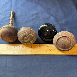 May include: Four door knobs are arranged in a row next to a yellow wooden ruler. Three knobs have a rusted, textured brown finish, with one featuring a floral pattern and a metal spindle. The third knob is smooth, glossy, and black in color.