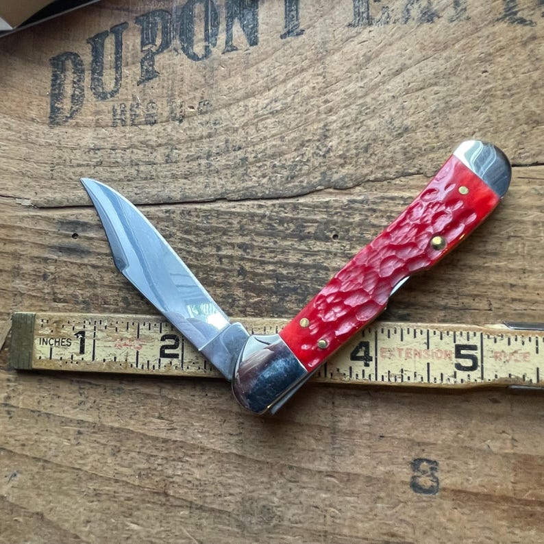 May include: A folding pocket knife with a single silver-colored steel blade and a textured, bright red handle. The knife is positioned over a yellow measuring tape, showing a total length of approximately 5.5 inches when fully extended.
