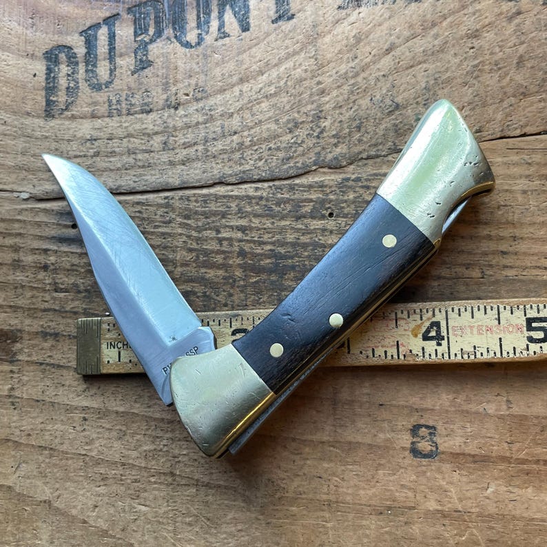 Puede incluir: Un cuchillo de bolsillo parcialmente abierto con una hoja plateada y un mango de madera oscura. El mango est&aacute; acentuado con lat&oacute;n y peque&ntilde;os remaches de lat&oacute;n. El cuchillo est&aacute; sobre una superficie de madera con una regla.