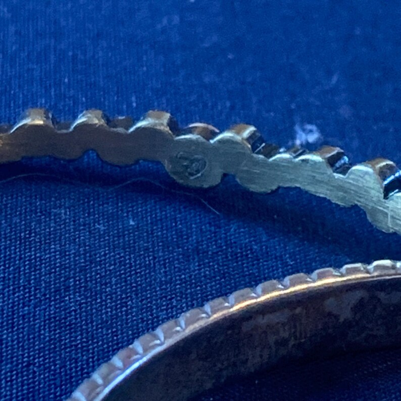 May include: Close-up of two silver-toned rings. The top ring has a scalloped edge with a small emblem. The bottom ring has a ridged edge. Both rings are set against a dark blue textured background.