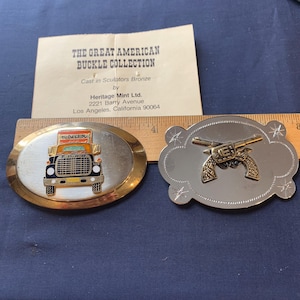 May include: Two belt buckles from The Great American Buckle Collection. One is oval with a truck design, the other is a silver-colored buckle with a gold-colored pistol design. The truck buckle has the word "KNOX" on it.