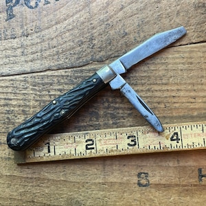 May include: A vintage pocket knife with a black textured handle and two open blades. The knife is positioned on a wooden surface next to a ruler marked in inches. The blades are silver and show signs of wear.