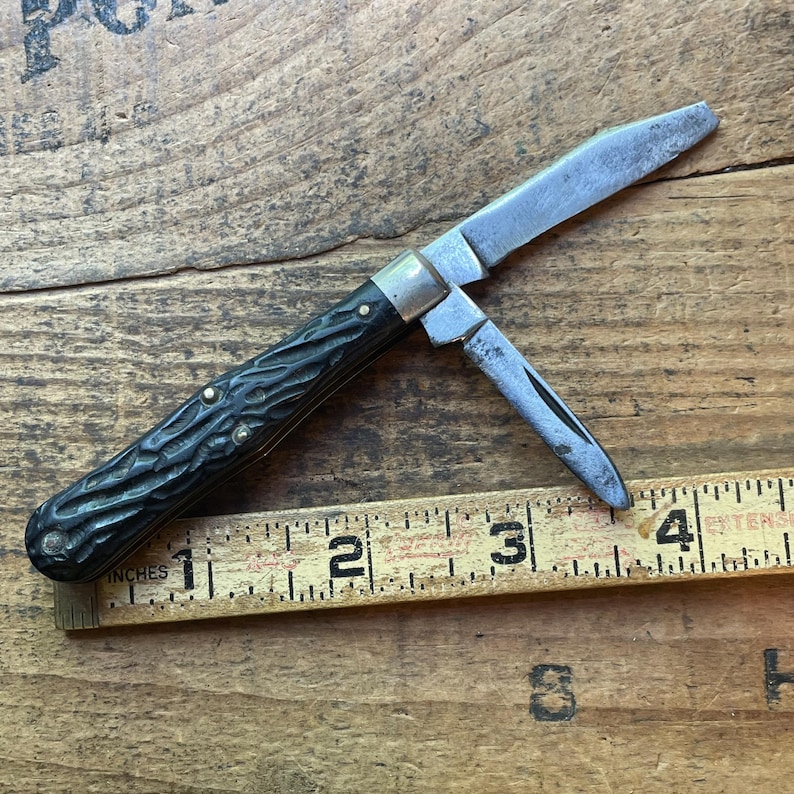 May include: A vintage pocket knife with a black textured handle and two open blades. The knife is positioned on a wooden surface next to a ruler marked in inches. The blades are silver and show signs of wear.