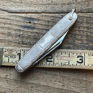 May include: A vintage silver pocket knife with an ornate, engraved design. The knife is approximately 3 inches long when closed. The blade is partially open, revealing a shiny, metallic edge. The knife is set against a wooden background.