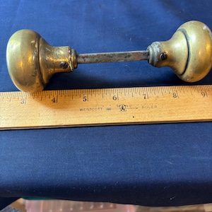 May include: A pair of round brass door knobs connected by a square metal spindle, positioned above a wooden Westcott ruler that shows the assembly spans approximately 6.5 inches in total length from the outer edge of one knob to the other.