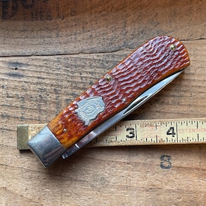 May include: A closed pocket knife with a textured, reddish-brown handle and a silver blade. The handle features a decorative silver emblem and brass rivets. A ruler is placed next to the knife.