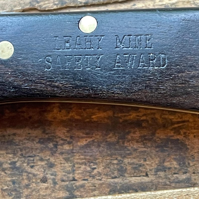 Puede incluir: Primer plano de un objeto de madera oscura con el texto grabado "LEAHY MINE SAFETY AWARD". El objeto tiene dos remaches de lat&oacute;n y un borde curvo. El fondo es una superficie de madera.