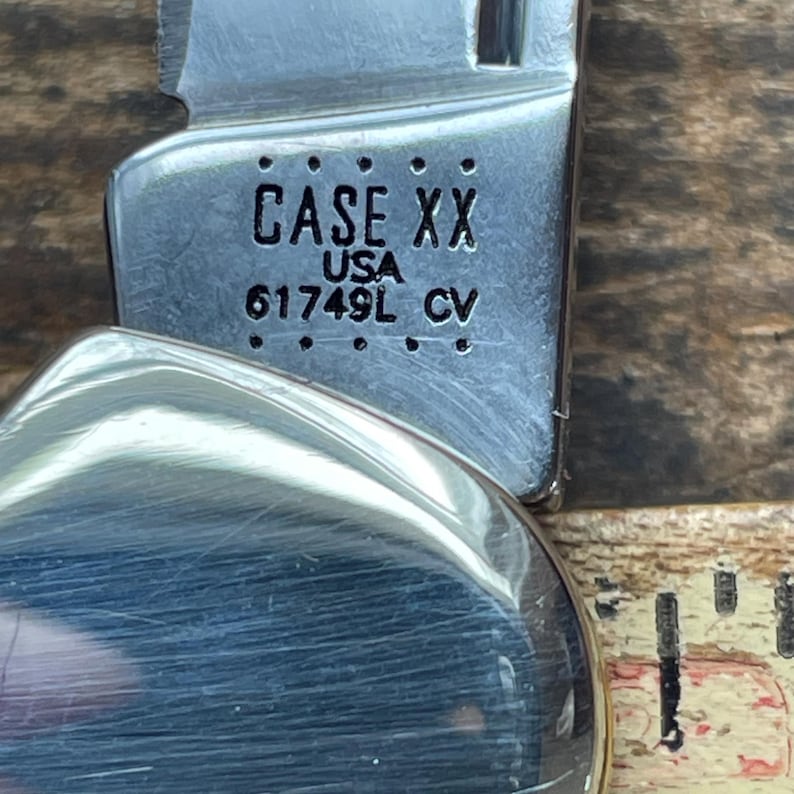 May include: A silver-colored metal pocket knife blade featuring the stamped text CASE XX USA 61749L CV. The blade is positioned next to a measuring tool, showing a portion of the scale with black markings on a light-colored background.