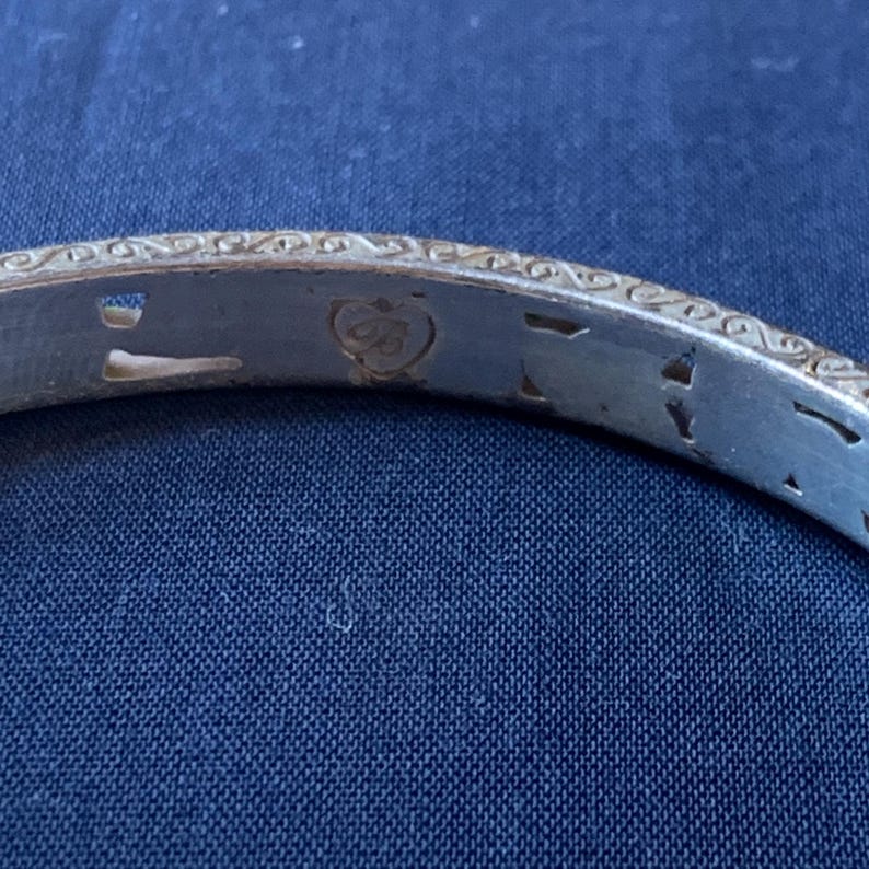 May include: A silver-toned bangle bracelet with an ornate, engraved design. The bracelet features a heart-shaped emblem with the letters "OB" inside. The background is a dark blue fabric.