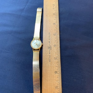 May include: A gold-toned wristwatch with a mesh band, next to a wooden ruler. The watch face is light-colored with gold hands and markers. The ruler shows measurements in inches.