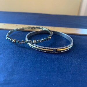 May include: Two silver bangles. One is adorned with small, colorful beads. The other has a gold-colored band in the center and a textured design. The bangles are displayed on a blue surface.