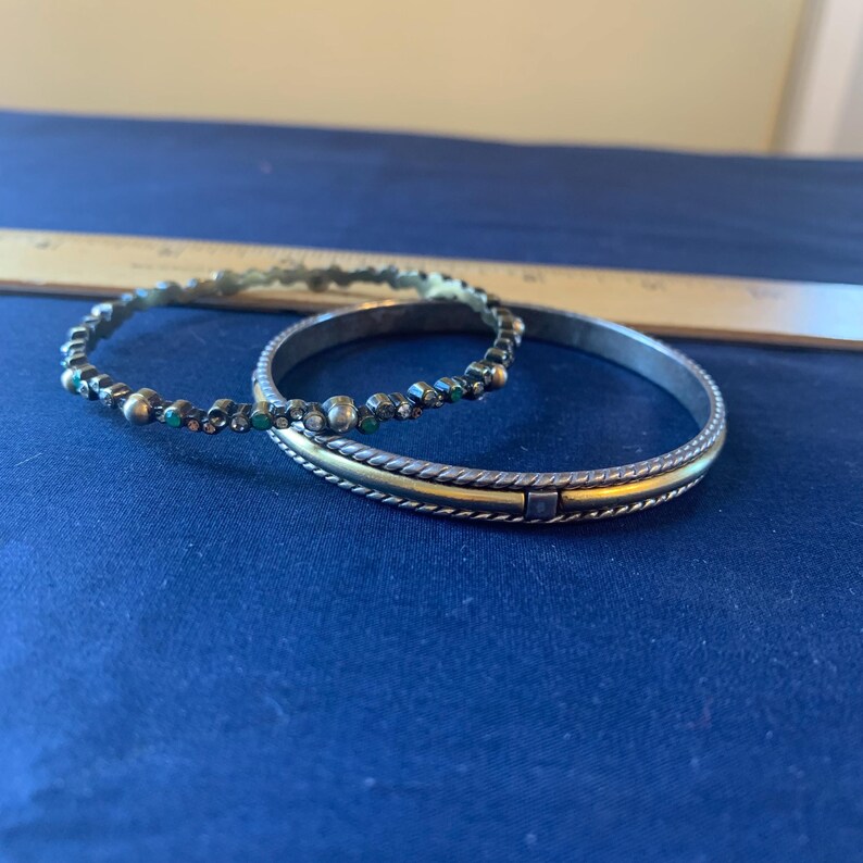 May include: Two silver bangles. One is adorned with small, colorful beads. The other has a gold-colored band in the center and a textured design. The bangles are displayed on a blue surface.