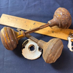 May include: Two antique metal door knobs with a rusted, textured finish and a central connecting spindle. A wooden ruler shows the assembly measures approximately 7.5 inches in length. Includes a circular white metal plate and a partial escutcheon.