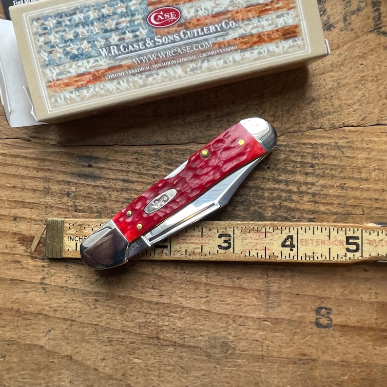 May include: A folding pocket knife with a textured red handle and silver metal bolsters, positioned above a yellow measuring tape showing a length of approximately 3.5 inches. The knife is accompanied by a box labeled W.R. Case & Sons Cutlery Co.
