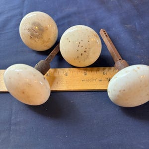 May include: Three round white ceramic door knobs with speckled textures and rusted metal spindles, positioned alongside a yellow wooden Westcott ruler that shows the spindles measure approximately 3 inches in length from the base of the knob.