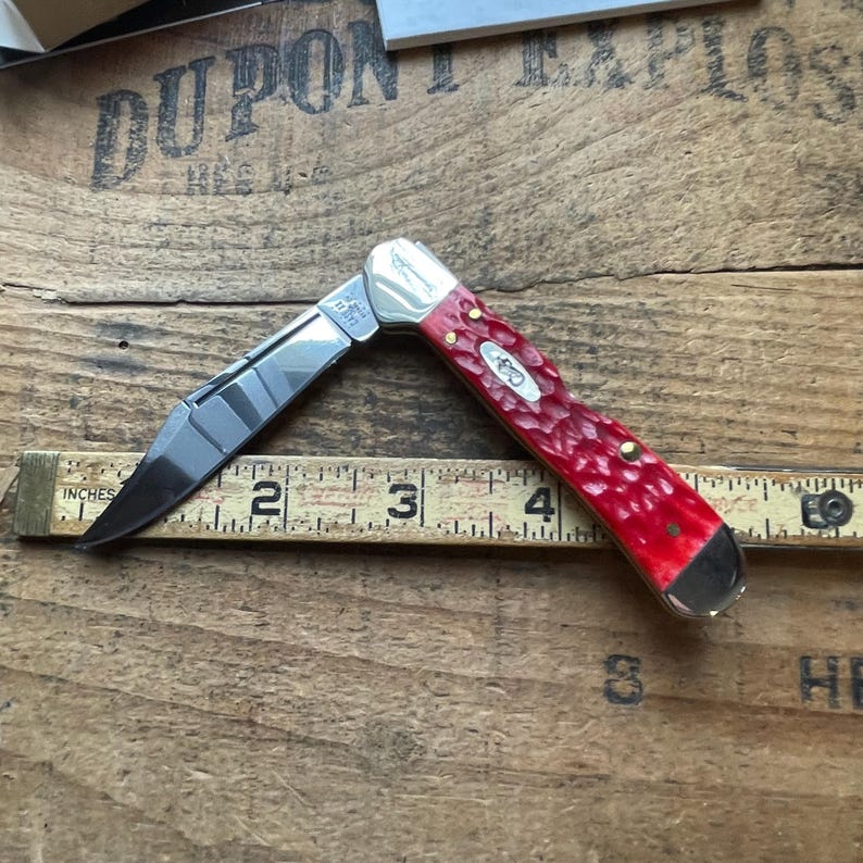 May include: A folding pocket knife with a polished silver blade and a textured red handle, positioned against a wooden measuring tape that shows the knife is approximately 4 inches long when partially open, featuring a silver bolster and rivets.