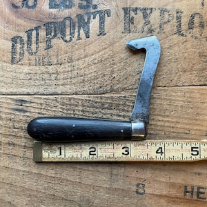 May include: Vintage metal knife with a curved blade and a black wooden handle. The knife is shown next to a ruler, measuring approximately 5 inches in length. The blade has a curved hook shape.