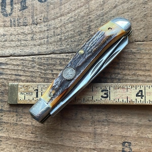 May include: A vintage pocket knife with a brown and cream handle, silver accents, and multiple blades. The knife features a decorative emblem and is shown next to a ruler with inch measurements.