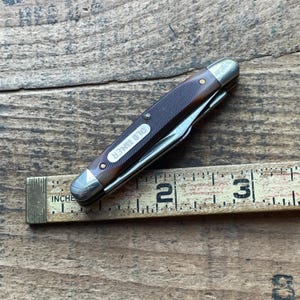 May include: A closed pocket knife with a brown handle and silver accents, labeled "OLD TIMER". The knife is next to a wooden ruler showing inches. The ruler measures approximately 3 inches.
