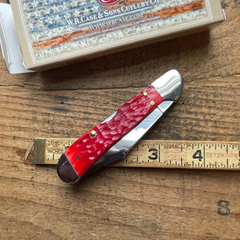 May include: A closed folding pocket knife with a textured red handle and polished silver metal bolsters. The knife is positioned over a wooden ruler showing a length of approximately 3.5 inches. A W.R. Case & Sons Cutlery Co. box is visible.